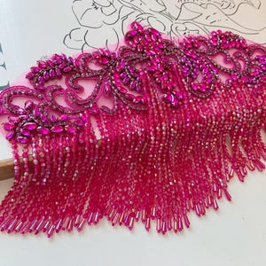 Puede incluir: Un vibrante adorno de cuentas fucsia con intrincados diseños florales y flecos colgantes. El adorno presenta una combinación de cristales grandes y facetados y cuentas más pequeñas, creando un adorno lujoso y llamativo.