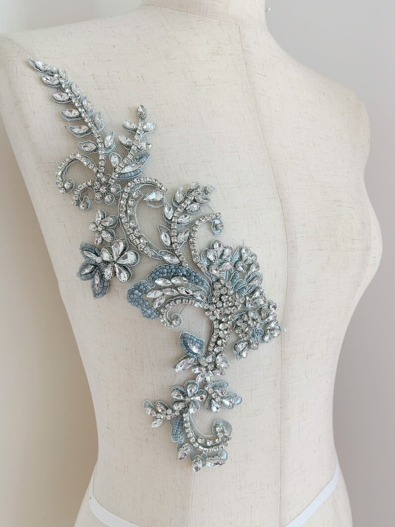 May include: A silver and blue beaded applique with a floral design. The applique is made with rhinestones and beads and is perfect for adding a touch of sparkle to any garment.