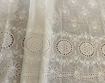 May include: White cotton fabric with a delicate eyelet embroidery pattern. The fabric features a repeating design of small flowers and circles. The edges of the fabric are scalloped.
