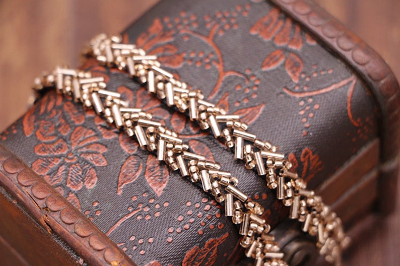 May include: Two gold chains with a chevron pattern, lying on a brown wooden box with a floral design.