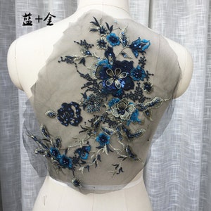 May include: A floral applique with intricate embroidery in shades of blue, navy, and gold. The design features detailed flowers, leaves, and beadwork on a sheer, gray fabric. The text "Blue + Gold" is visible in the upper left corner.