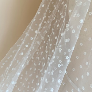Pode incluir: Tecido branco transparente com um delicado padrão floral. O tecido é macio e leve, tornando-o ideal para projetos de artesanato e costura.