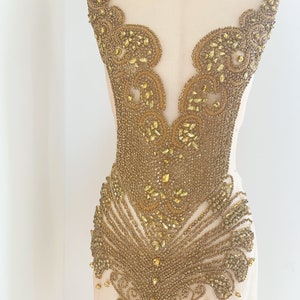 May include: Gold beaded and sequined applique with a delicate, intricate design. The applique features a V-shaped neckline and a flowing, cascading pattern.