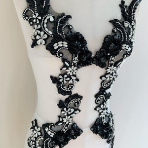 Peut inclure: Applique en dentelle noire avec des motifs floraux et de feuilles, ornée de strass transparents. L'applique est présentée sur un mannequin, mettant en valeur ses détails complexes, idéale pour embellir des vêtements.