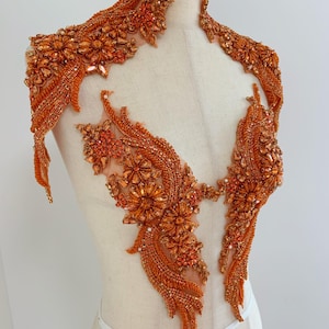 May include: An orange beaded applique, possibly for clothing decoration. The design includes detailed floral and flowing patterns, with a combination of beads and rhinestones. The applique is shown on a mannequin, highlighting its shape and form.