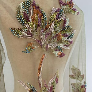 Puede incluir: Una prenda de tela de malla verde oliva transparente con intrincados bordados florales. El diseño está adornado con lentejuelas y cuentas de vidrio multicolores en tonos rosa, dorado, bronce y azul iridiscente, formando grandes motivos botánicos.