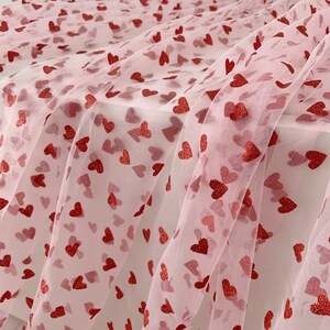 May include: Pink tulle fabric with a red heart pattern. The hearts are scattered randomly across the fabric.