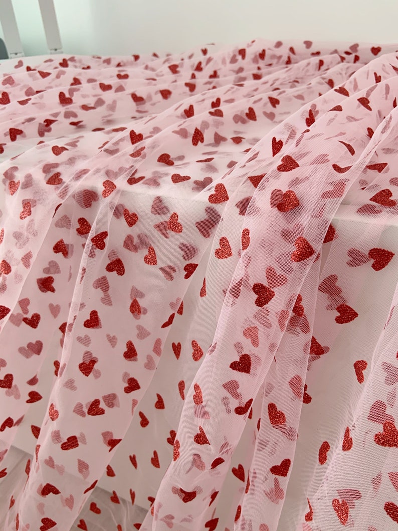May include: Pink tulle fabric with a red heart pattern. The hearts are scattered randomly across the fabric.