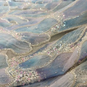 Op de afbeelding: Close-up van een stof met een vloeiend, golfachtig ontwerp. De stof heeft een verloop van blauw, paars en wit, versierd met pailletten en kleine, roze kralen. De randen zijn omlijnd met zilveren kralen.