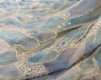 May include: Close-up of a fabric with a flowing, wave-like design. The fabric features a gradient of blue, purple, and white, embellished with sequins and small, pink beads. The edges are outlined with silver beading.