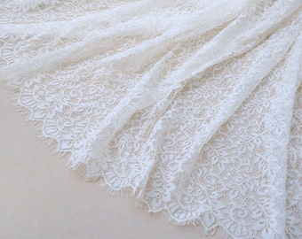May include: Ivory-colored lace fabric with a delicate floral pattern. The fabric is draped, showcasing its intricate design and scalloped edges. This textile is suitable for bridal wear, dresses, or other craft projects.