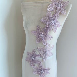 May include: A cascade of lavender floral appliques with beaded centers and sequin accents. The delicate, sheer fabric is draped over a mannequin, showcasing the intricate design and texture of the embellishments.