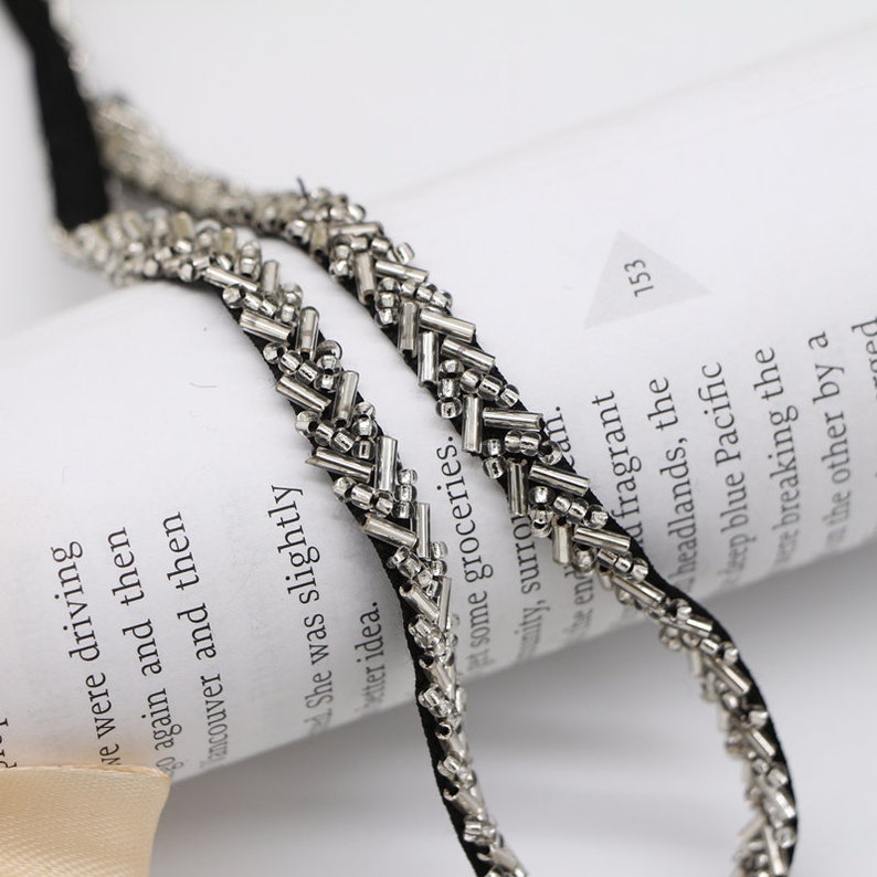 May include: Two strands of silver beaded trim on black ribbon. The trim is made of small, silver beads and metal bars arranged in a chevron pattern.