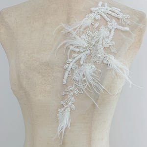 May include: An elongated white applique featuring intricate beadwork, sequins, and soft feathers attached to a sheer mesh backing. The design consists of floral and vine-like patterns created with white beads and pearls, accented by delicate white feathers.
