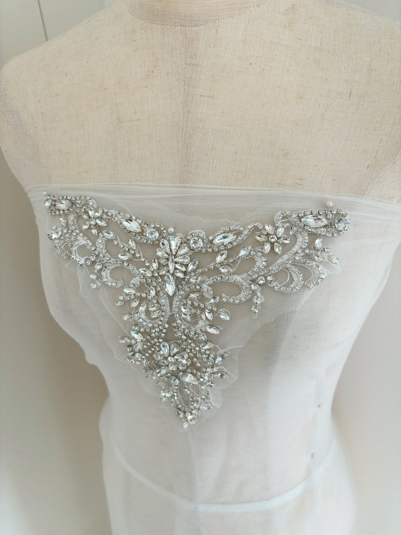 May include: A white tulle fabric with a large, ornate, silver rhinestone embellishment. The embellishment is in the shape of a teardrop and features a variety of sizes and shapes of rhinestones.