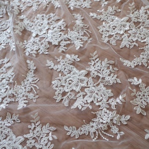 May include: White lace fabric with a delicate floral pattern. The lace is sheer and has a soft, flowing texture.