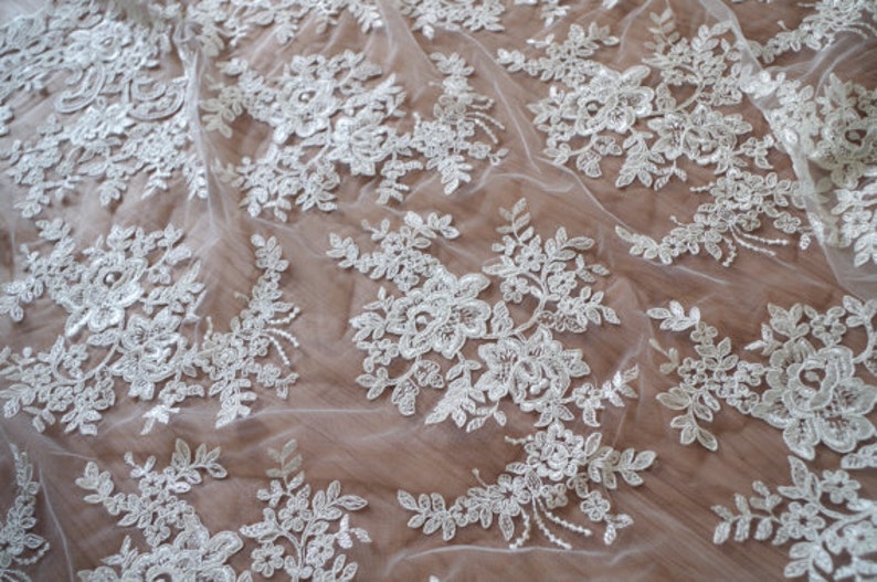 May include: White lace fabric with a delicate floral pattern. The lace is sheer and has a soft, flowing texture.