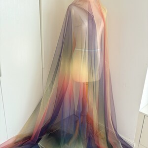 May include: Rainbow ombre tulle fabric. The fabric is sheer and has a gradient of colors, starting with yellow at the top and transitioning to red, purple, and green at the bottom.