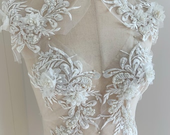 May include: White lace applique with pearl embellishments. The lace features floral designs and delicate beading. This applique is perfect for adding a touch of elegance to any garment.