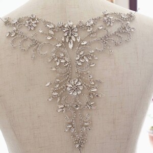 May include: A silver rhinestone applique with a floral design. The applique is on a white fabric background.