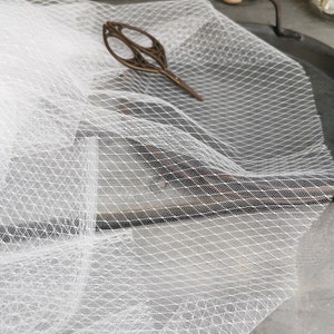 Puede incluir: Tela de tul blanco con un fino patrón de malla en forma de diamante. La tela está drapeada sobre una superficie metálica.