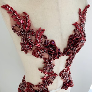 Könnte beinhalten: Rote, aufwendig gearbeitete Applikation mit floralen und Blattmotiven. Die Applikation ist mit verschiedenen roten Perlen und Pailletten verziert, die einen schimmernden Effekt erzeugen. Sie ist auf einem transparenten Mesh-Träger angebracht.