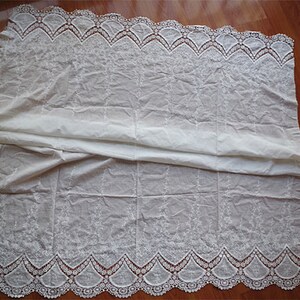 May include: White cotton fabric with a delicate embroidered design and a scalloped lace trim. The fabric is folded in half, showing both sides of the lace.