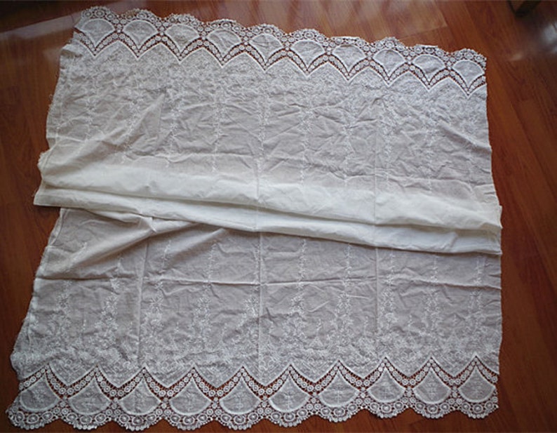 May include: White cotton fabric with a delicate embroidered design and a scalloped lace trim. The fabric is folded in half, showing both sides of the lace.