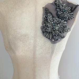 May include: A decorative floral appliqué featuring intricate silver and dark grey beadwork on a sheer grey mesh fabric. The design consists of two distinct flower shapes with detailed petal textures, secured with small white pins to a mannequin.