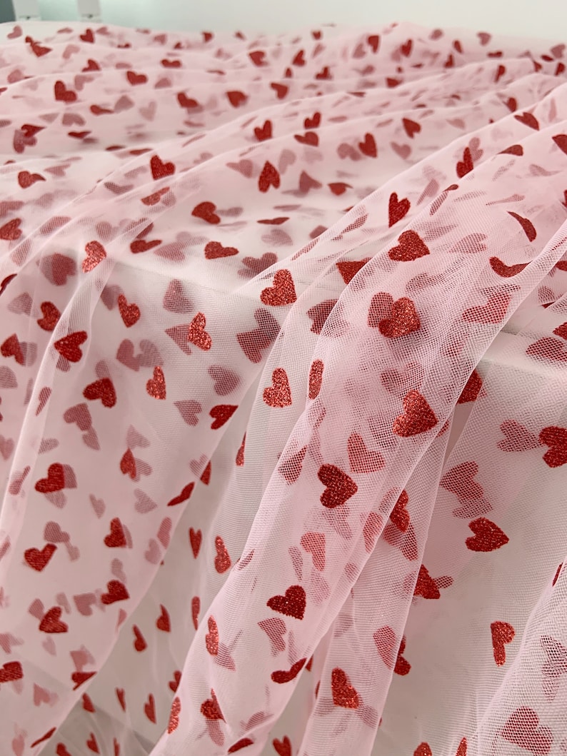 May include: Pink tulle fabric with red glitter hearts printed on it. The fabric is sheer and delicate, and the hearts are scattered randomly across the surface.