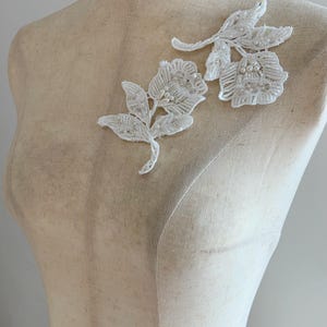 May include: Two white floral lace appliqués featuring intricate embroidery and small white pearl bead embellishments. Each piece depicts a flower with leaves and stems, crafted from white thread with a textured finish, displayed on a mannequin.