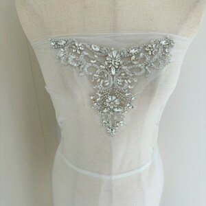 May include: A white sheer fabric with a large, ornate, crystal embellishment. The embellishment is in the shape of a V and features a variety of sizes and shapes of crystals.