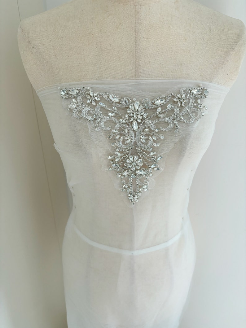 May include: A white sheer fabric with a large, ornate, crystal embellishment. The embellishment is in the shape of a V and features a variety of sizes and shapes of crystals.