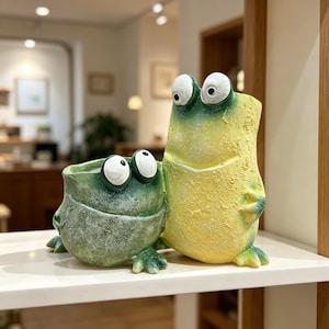 May include: A pair of frog-shaped planters featuring a textured finish. The smaller planter is dark green with two prominent white eyes, while the taller, upright planter is yellow with a green top section and two large white eyes on top.