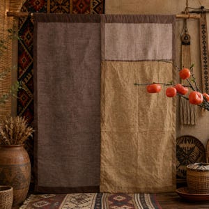 Op de afbeelding: Een rechthoekige wandhanger met een donkerbruin linnen paneel aan de linkerkant en een tweekleurig lichtbruin en beige linnen paneel aan de rechterkant, opgehangen aan een houten roede. Een tak met verschillende feloranje kaki vruchten.