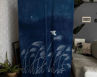 Pode incluir: Uma cortina retangular de tecido azul índigo com um design serigrafado em branco de ervas altas que balançam, uma pequena lua crescente no canto superior esquerdo e um único pássaro em voo. A cortina está pendurada numa vara de bambu.