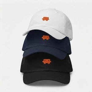 May include: A stack of three baseball caps in white, navy blue, and black. Each cap features a small, orange, pixelated, rectangular character embroidered on the front center. The hats have a structured crown and a curved brim.