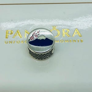 May include: A round silver charm featuring a dark blue mountain silhouette with a white peak and a branch of pink cherry blossoms. The sides of the charm are encrusted with small clear stones, and the piece is set against a white Pandora box.
