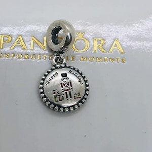 May include: A silver dangle charm featuring a circular disc with a beaded edge. The center displays a colorful nutcracker figure and wrapped gifts with the text Happy Holidays engraved in black script. The charm has a large, smooth bail at the top.