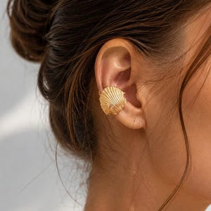 May include: A gold-toned ear cuff shaped like a small, textured seashell. The piece features detailed, radiating ridges across its surface, mimicking the natural form of a scallop shell, and is designed to sit securely along the outer edge of the ear.