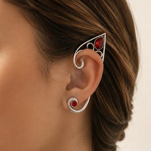 May include: A silver-toned ear cuff and matching stud earring set featuring a swirling metal design. The ear cuff has a pointed top section with a teardrop-shaped red stone, while the stud earring features a smaller circular red stone.