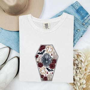 May include: A white t-shirt featuring a central graphic of a coffin shape filled with illustrations of a black spider, dark red roses, beige vampire fangs, grey candles, and small black bats. The inner neck label reads Comfort Colors.