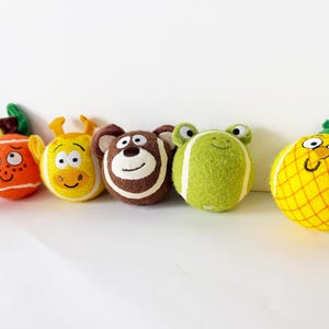 Könnte beinhalten: Eine Sammlung von sechs runden Plüschspielzeugen, die verschiedenen Charakteren nachempfunden sind. Das Set enthält eine violette Aubergine, eine orangefarbene Frucht, eine gelbe Giraffe, einen braunen Bären, einen grünen Frosch und eine gelbe Ananas.