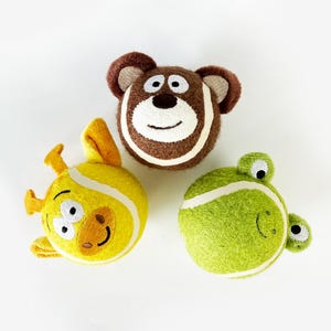 Pode incluir: Três brinquedos de pelúcia redondos em forma de bolas de ténis, com rostos de animais bordados. Um urso castanho, uma girafa amarela com orelhas pequenas e um sapo verde com olhos salientes, todos com detalhes em feltro branco.