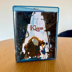 Puede incluir: Una caja de Blu-ray azul con la película de animación Klaus. La portada muestra a una figura central barbuda con abrigo marrón, una persona con abrigo azul y un niño pequeño, con el texto Welcome To The Jingle y el título Klaus en rojo.