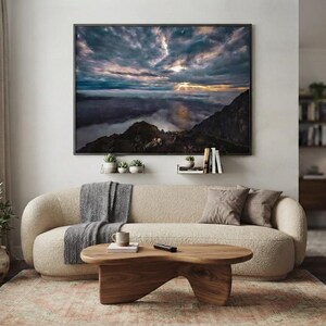 Könnte beinhalten: Ein großes rechteckiges Wandbild mit einer dramatischen Bergkette über einer dichten Wolkendecke bei Sonnenuntergang, mit dunklen Felsgipfeln, einem tiefblauen Himmel und goldenem Licht, das durch die Wolken bricht, platziert über einem beigen Sofa.
