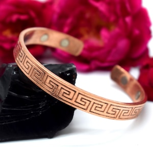 May include: A copper cuff bracelet featuring a repeating Greek key pattern engraved along the outer surface. The bracelet has an open design with two small circular magnets visible on the interior ends of the metal band.