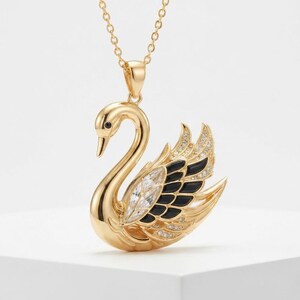 Può includere: Una collana con ciondolo color oro a forma di cigno. Il cigno presenta un corpo in oro lucido, dettagli in smalto nero sulle ali e decorazioni in cristallo trasparente lungo l'ala e il collo. È appeso a una sottile catenina color oro.