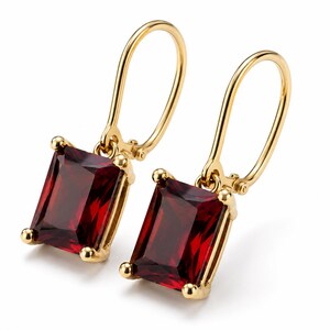 May include: A pair of gold drop earrings featuring rectangular, deep red faceted gemstones held by four prongs. The earrings have a polished gold finish and a secure lever-back closure mechanism, showcasing a classic design with vibrant red stones.