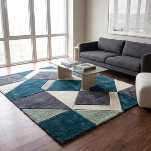 Puede incluir: Una alfombra rectangular grande con un patrón geométrico de bloques en color verde azulado, gris carbón, azul claro y crema. La alfombra está colocada sobre un suelo de madera oscura en una habitación con un sofá gris oscuro y un sillón blanco.
